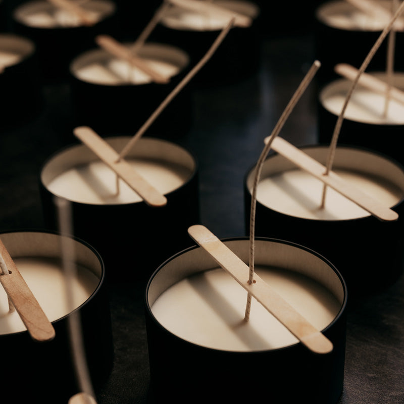 A moody black photo with several candles with white wax cooling.  They all hav a Popsicle stick across the candle tin to help set the candle's wick during the cooling process.